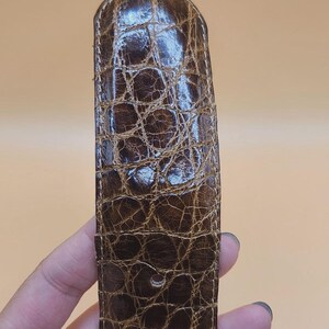 May include: Close-up of a brown leather belt with a crocodile skin texture. The belt has a rectangular shape with rounded edges and several holes for adjustment. The leather has a glossy finish.