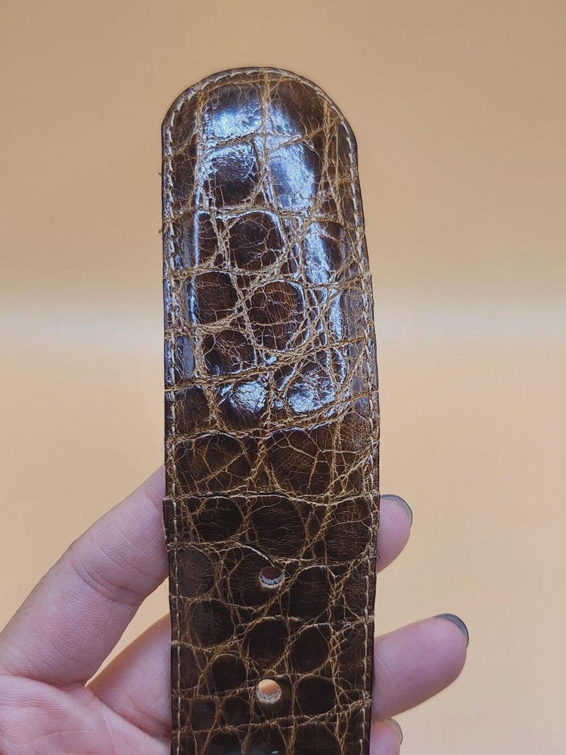 May include: Close-up of a brown leather belt with a crocodile skin texture. The belt has a rectangular shape with rounded edges and several holes for adjustment. The leather has a glossy finish.