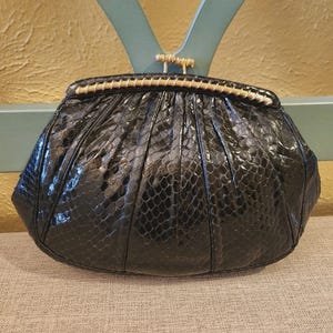 Op de afbeelding: Een zwarte, getextureerde clutch met een goudkleurige sluiting en afwerking. De tas heeft een afgeronde vorm met plooien die van bovenaf uitstralen. De sluiting heeft een rechthoekige vorm met een goudkleurige balk.