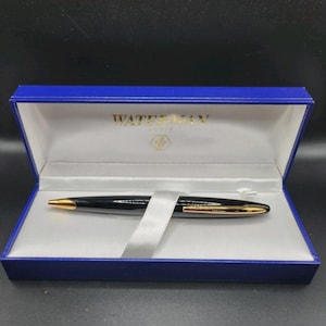 Könnte beinhalten: Ein schwarzer Waterman-Stift mit goldfarbenen Akzenten liegt in einer blauen Präsentationsbox. Die Box hat ein weißes Innenfutter und ein Satinband. Das Waterman-Logo ist auf der Box zu sehen.