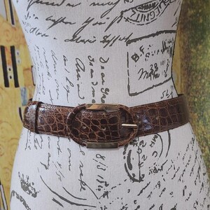 May include: A brown belt with a faux crocodile skin texture is fastened around a mannequin. The belt has a gold-colored oval buckle. The belt is a medium brown color.