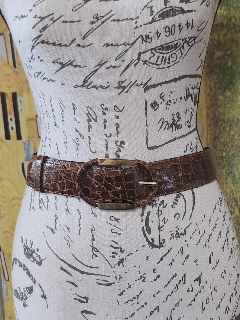 May include: A brown belt with a faux crocodile skin texture is fastened around a mannequin. The belt has a gold-colored oval buckle. The belt is a medium brown color.