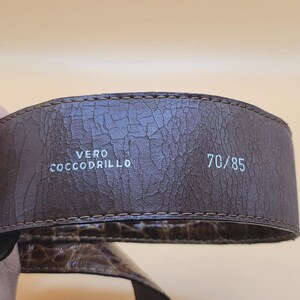 May include: Close-up of a brown leather belt with a textured surface. The belt has the text "VERO COCCODRILLO" and the size "70/85" printed in white. The belt is likely made of crocodile leather.