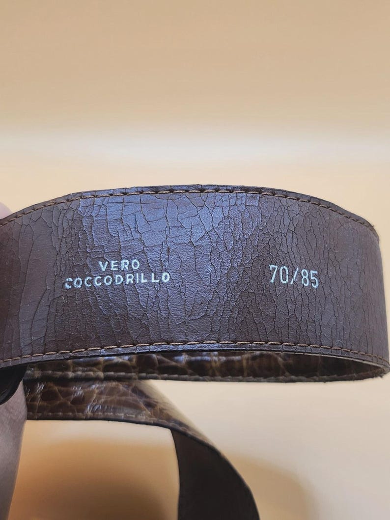 May include: Close-up of a brown leather belt with a textured surface. The belt has the text "VERO COCCODRILLO" and the size "70/85" printed in white. The belt is likely made of crocodile leather.