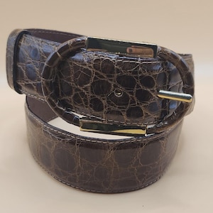 May include: A brown belt with a faux crocodile skin texture. The belt features a large, oval-shaped buckle with gold-colored metal accents. The belt is rolled up, showcasing its design and construction.