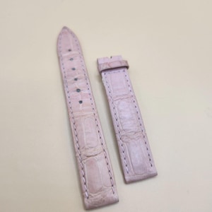 May include: A two-piece light pink watch strap featuring a textured alligator-style embossed pattern. The longer piece includes a pointed tip and multiple adjustment holes, while the shorter piece features a matching sliding keeper loop.