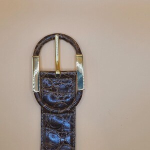 May include: A brown leather belt with a gold-colored buckle. The belt has a textured, crocodile-like pattern. The buckle is a gold-colored oval shape with a gold-colored bar in the center. The belt is a classic accessory.