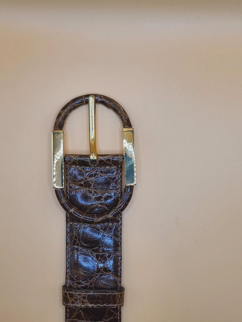 May include: A brown leather belt with a gold-colored buckle. The belt has a textured, crocodile-like pattern. The buckle is a gold-colored oval shape with a gold-colored bar in the center. The belt is a classic accessory.