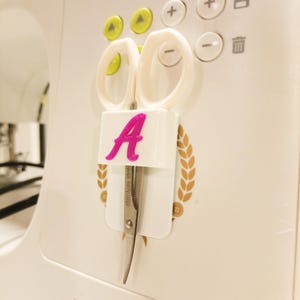 Op de afbeelding: Witte naaimachine met een schaar die op zijn plaats wordt gehouden door een witte houder. De houder heeft een roze letter "A" erop. De schaar is van roestvrij staal.