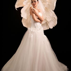 May include: A white bridal gown with a tulle skirt and a fitted, floral-embroidered bodice. The bride wears a veil and necklace. A large paper flower backdrop is behind the bride, set against a black background.