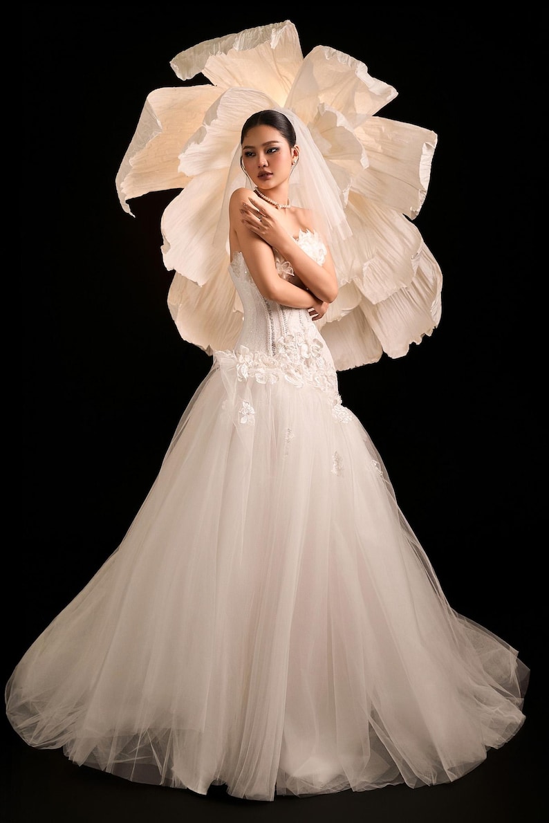 May include: A white bridal gown with a tulle skirt and a fitted, floral-embroidered bodice. The bride wears a veil and necklace. A large paper flower backdrop is behind the bride, set against a black background.
