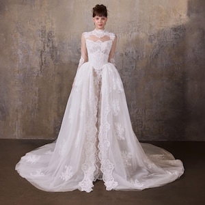 Könnte beinhalten: Ein bodenlanges weißes Kleid mit hohem Ausschnitt, langen transparenten Ärmeln und einem Korsett-Oberteil. Das Kleid ist mit aufwendigen Spitzenapplikationen und einem mehrlagigen Tüllrock versehen, der vorne offen ist und einen Unterrock zeigt.