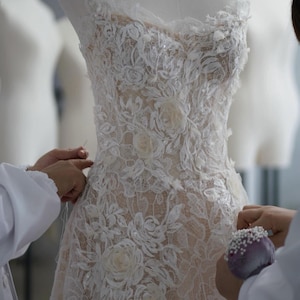 May include: A strapless gown featuring intricate white floral lace overlays on a nude-colored base. The bodice and skirt are adorned with three-dimensional fabric flower appliqu&eacute;s and subtle sequin embellishments throughout the lace pattern.