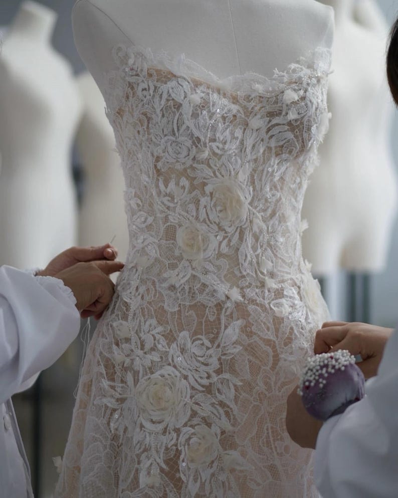 May include: A strapless gown featuring intricate white floral lace overlays on a nude-colored base. The bodice and skirt are adorned with three-dimensional fabric flower appliqu&eacute;s and subtle sequin embellishments throughout the lace pattern.