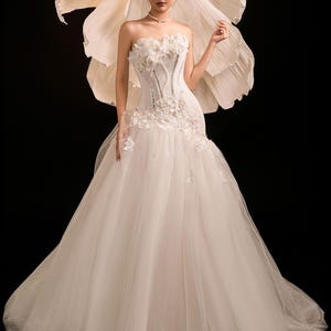 May include: A white wedding dress with a strapless bodice adorned with floral details and beadwork. The skirt is made of tulle. A large paper flower is behind the dress, and a veil is worn. The dress is floor-length.