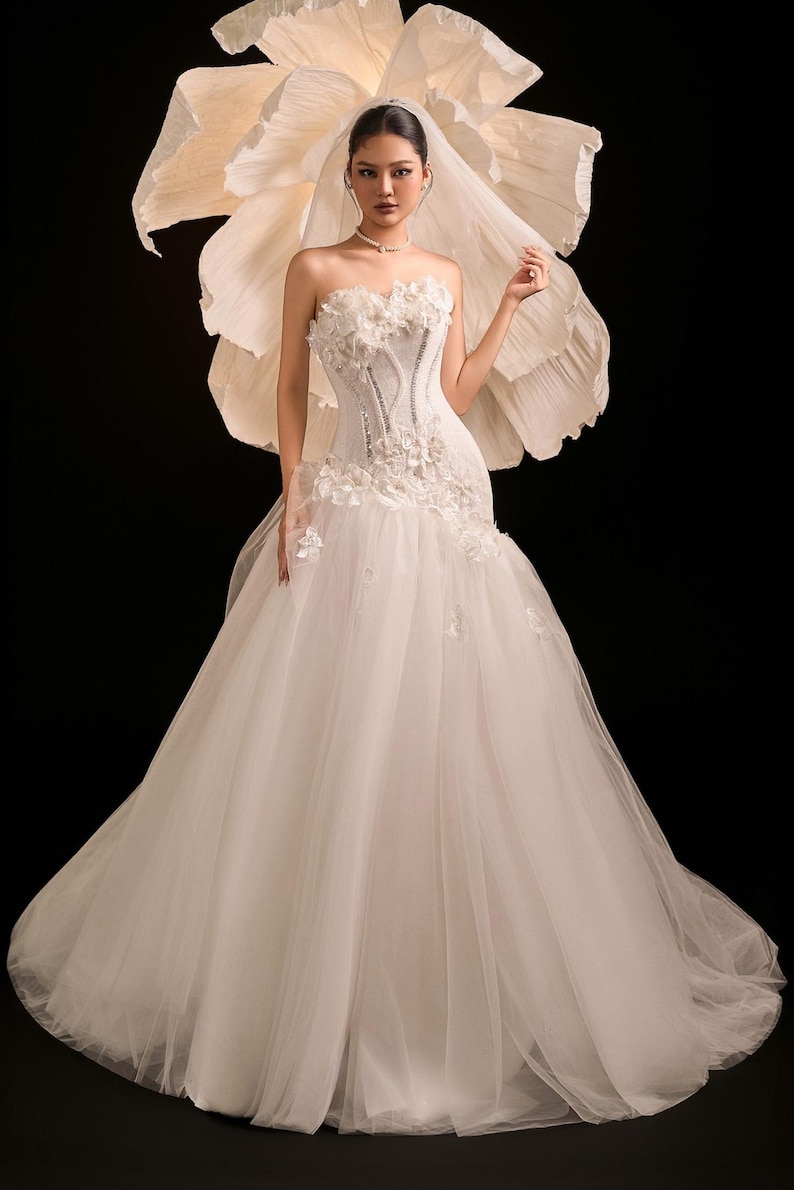 May include: A white wedding dress with a strapless bodice adorned with floral details and beadwork. The skirt is made of tulle. A large paper flower is behind the dress, and a veil is worn. The dress is floor-length.