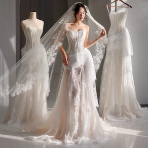 May include: A white wedding dress with a corset bodice and a flowing skirt, adorned with delicate lace details. A bride wears the dress, holding a veil. Two additional white wedding dresses are displayed on mannequins, showcasing the design.