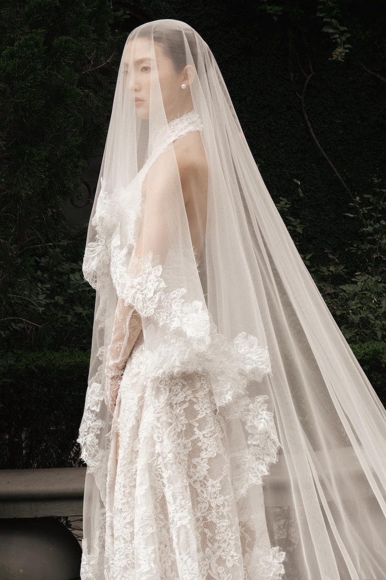 May include: A white lace wedding dress with a high neckline and long veil. The dress features intricate floral lace detailing and a flowing skirt. The veil cascades down the back, creating a romantic and elegant look.
