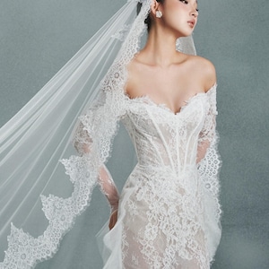 May include: A white, off-the-shoulder wedding dress with intricate lace detailing. The dress features a fitted bodice and a flowing skirt. A matching lace veil cascades down the back. The overall style is elegant and romantic.