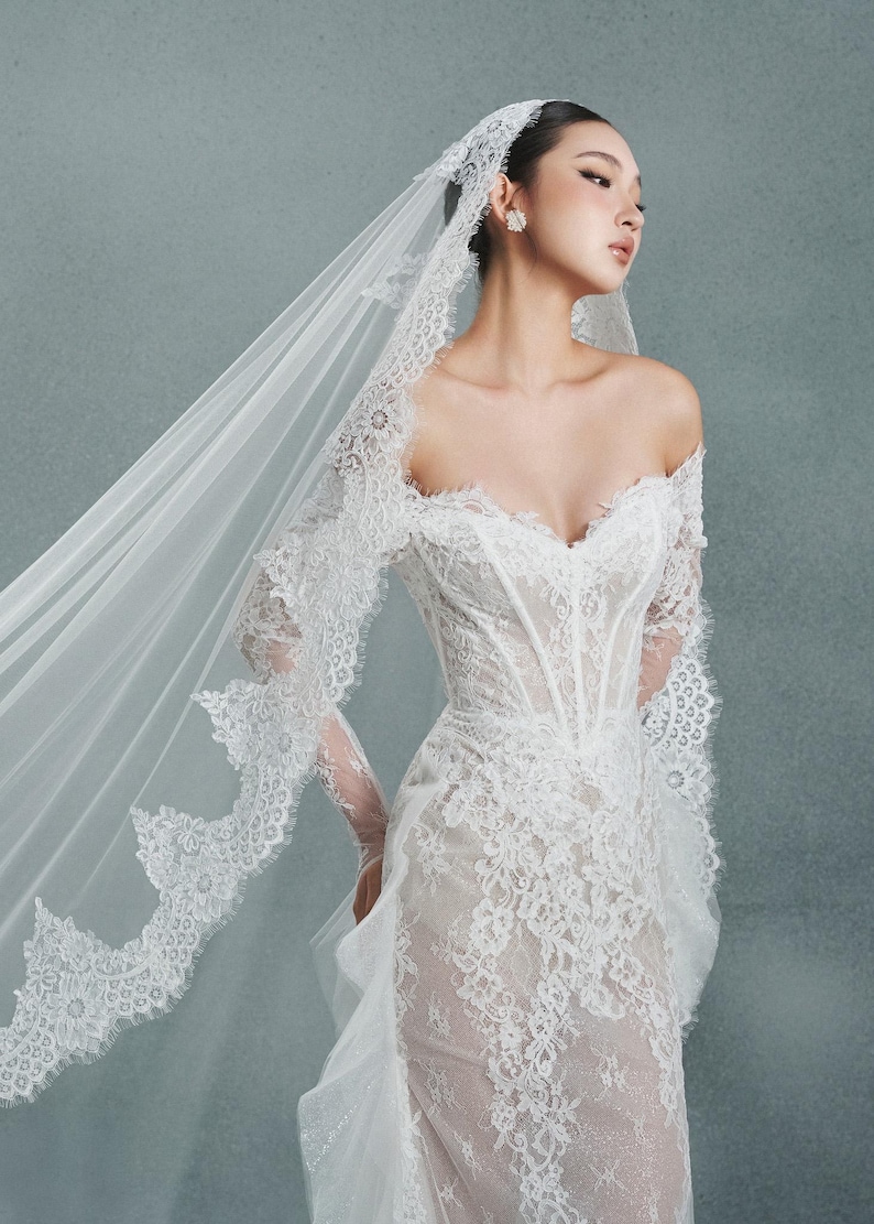 May include: A white, off-the-shoulder wedding dress with intricate lace detailing. The dress features a fitted bodice and a flowing skirt. A matching lace veil cascades down the back. The overall style is elegant and romantic.