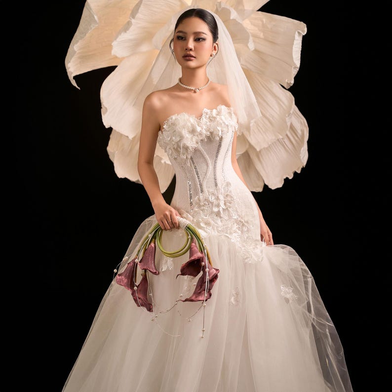 May include: A white strapless wedding dress with floral detailing and a tulle skirt. The bride carries a bouquet of burgundy calla lilies. A large paper flower is behind her, and she wears a veil and a pearl necklace.