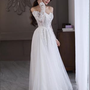 May include: A white off-the-shoulder wedding gown with long, sheer sleeves. The bodice is embellished with intricate beadwork and sequins, and the skirt flows to the floor. The dress is made of delicate, lightweight fabric, creating an elegant silhouette.