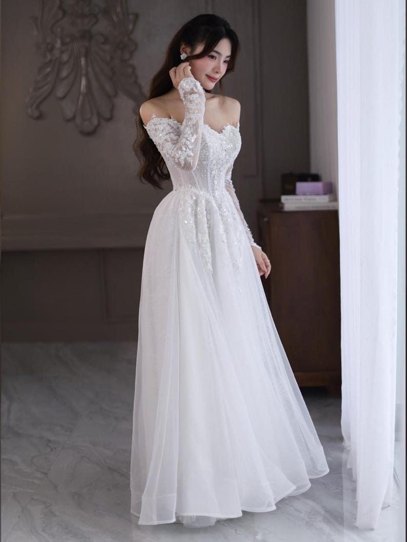 May include: A white off-the-shoulder wedding gown with long, sheer sleeves. The bodice is embellished with intricate beadwork and sequins, and the skirt flows to the floor. The dress is made of delicate, lightweight fabric, creating an elegant silhouette.