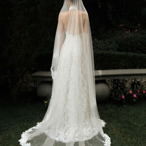 May include: A white lace wedding dress with a long train and a sheer veil. The dress has a halter neckline and a fitted bodice. The veil cascades down the back, and the train is trimmed with lace. The dress is worn outdoors on a grassy area.