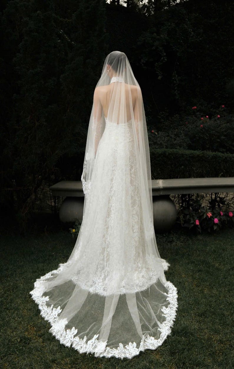 May include: A white lace wedding dress with a long train and a sheer veil. The dress has a halter neckline and a fitted bodice. The veil cascades down the back, and the train is trimmed with lace. The dress is worn outdoors on a grassy area.