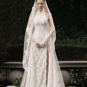 May include: A white lace wedding dress with a high neckline and long sleeves. The dress features a thigh-high slit and is paired with a long veil. The intricate floral lace design covers the entire gown, creating an elegant and romantic look.