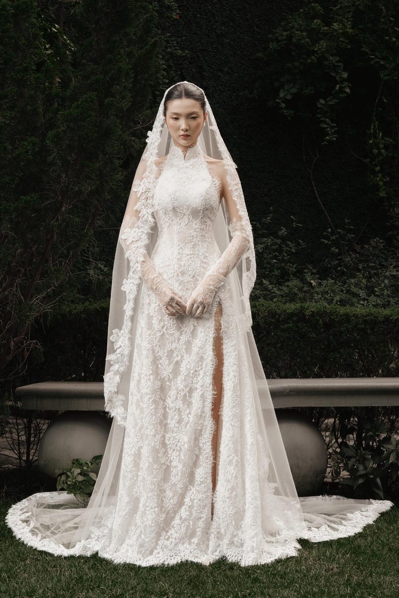 May include: A white lace wedding dress with a high neckline and long sleeves. The dress features a thigh-high slit and is paired with a long veil. The intricate floral lace design covers the entire gown, creating an elegant and romantic look.