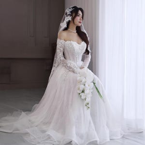 May include: A white off-the-shoulder wedding dress with long lace sleeves and a full skirt. The bodice is embellished with sequins and beads. A white veil and a bouquet of white flowers complete the bridal look.