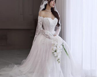 Pode incluir: Vestido de noiva branco com decote ombro a ombro, mangas compridas de renda e saia ampla. O corpete é adornado com lantejoulas e contas. Um véu branco e um buquê de flores brancas completam o visual.