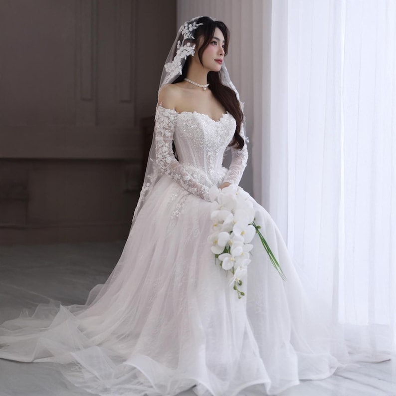 May include: A white off-the-shoulder wedding dress with long lace sleeves and a full skirt. The bodice is embellished with sequins and beads. A white veil and a bouquet of white flowers complete the bridal look.