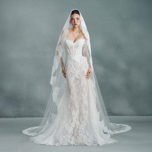 May include: A white lace wedding dress with a sweetheart neckline and long sleeves. The dress features intricate lace detailing and a fitted bodice that flares into a mermaid silhouette. A matching lace veil drapes over the shoulders.