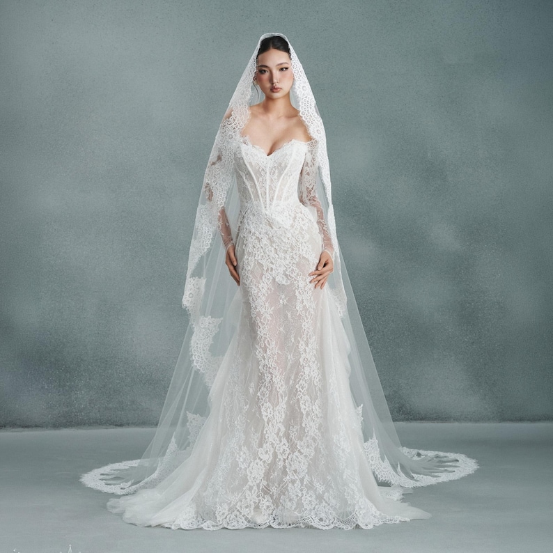 May include: A white lace wedding dress with a sweetheart neckline and long sleeves. The dress features intricate lace detailing and a fitted bodice that flares into a mermaid silhouette. A matching lace veil drapes over the shoulders.