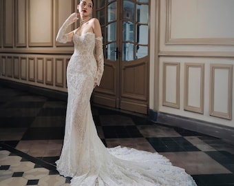May include: A white, strapless wedding gown with intricate lace detailing. The gown features a fitted bodice, long sleeves, and a flowing train. The bride is wearing a pearl necklace and matching gloves. The setting is a hallway with a checkered floor.