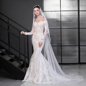 May include: A white, off-the-shoulder wedding dress with long sleeves and a mermaid silhouette. The dress features detailed lace. A matching veil drapes from the head, extending into a long train. The bride is standing near a black staircase.