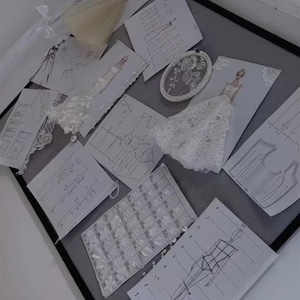 May include: A collection of fashion design sketches and sewing patterns arranged on a board. The display includes white paper templates with black line drawings of garment silhouettes, fabric swatches with floral lace, and an oval embroidery hoop.