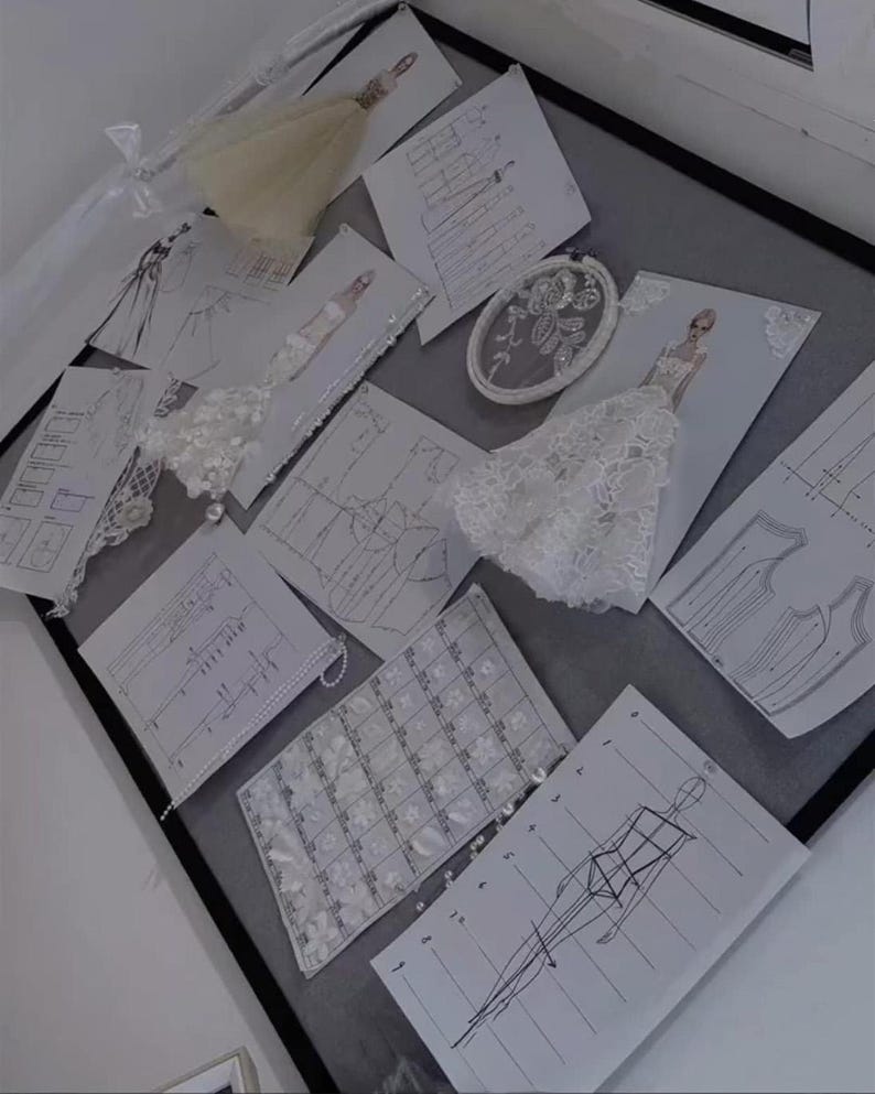 May include: A collection of fashion design sketches and sewing patterns arranged on a board. The display includes white paper templates with black line drawings of garment silhouettes, fabric swatches with floral lace, and an oval embroidery hoop.