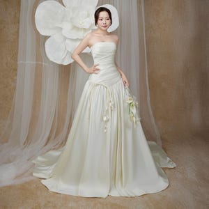 May include: A strapless ivory wedding gown with a flowing skirt and a slight train. The bodice features delicate ruching and a decorative waist detail. The bride is holding a bouquet of white flowers. A large white paper flower is in the background.