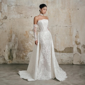 May include: A white strapless floor-length wedding dress featuring intricate lace embroidery, a corset-style bodice, and a detachable overskirt. The garment includes matching sheer lace sleeves that extend to the wrists and a high lace collar.