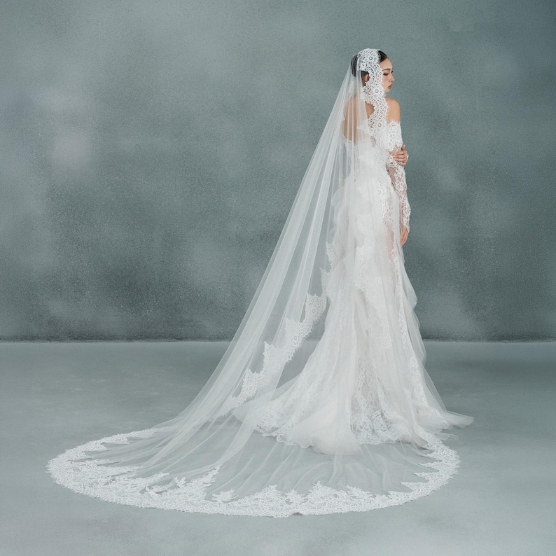 May include: A white wedding dress with a long, flowing train and a matching veil. The veil and dress are adorned with delicate lace detailing. The bride is standing with her arms crossed.