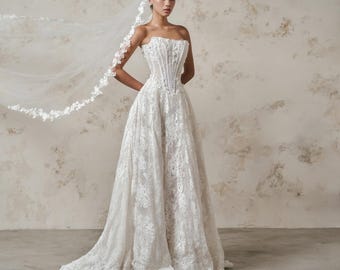 Pode incluir: Um vestido de noiva branco sem alças com um corpete tipo espartilho estruturado com barbatanas verticais e uma saia comprida coberta por intrincados bordados de renda floral. Um véu longo e transparente a condizer com rebordo de renda.