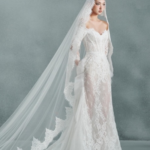 May include: Elegant white lace wedding gown with long sleeves and a flowing veil. The dress features a sweetheart neckline and intricate floral lace detailing. The veil cascades down the back, creating a romantic and ethereal look.