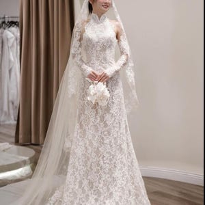 May include: A white lace wedding dress with long sleeves and a high neckline. The dress features intricate floral lace detailing and a flowing skirt. A sheer veil and a bouquet of white flowers complete the bridal look.