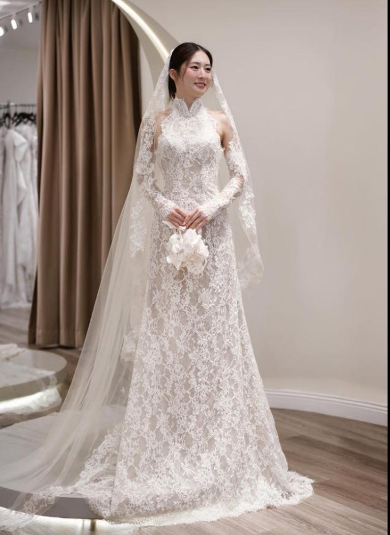 May include: A white lace wedding dress with long sleeves and a high neckline. The dress features intricate floral lace detailing and a flowing skirt. A sheer veil and a bouquet of white flowers complete the bridal look.