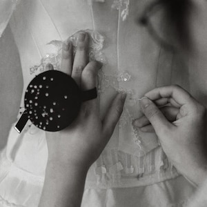 May include: A white garment featuring intricate lace appliqu&eacute; and sequin detailing. A round black pin cushion with numerous silver pins is attached to the wrist, while a needle is used to secure the delicate lace fabric onto the white base.