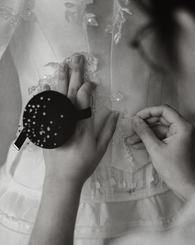 May include: A white garment featuring intricate lace appliqu&eacute; and sequin detailing. A round black pin cushion with numerous silver pins is attached to the wrist, while a needle is used to secure the delicate lace fabric onto the white base.