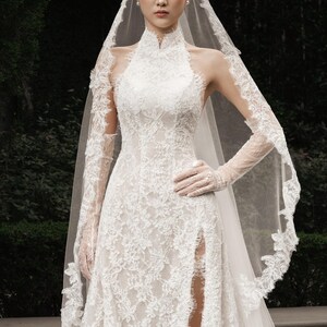 May include: A white lace wedding dress with a high neckline and a thigh-high slit. The dress features long, sheer sleeves and a matching veil with lace trim. The bride is wearing white gloves and pearl earrings.